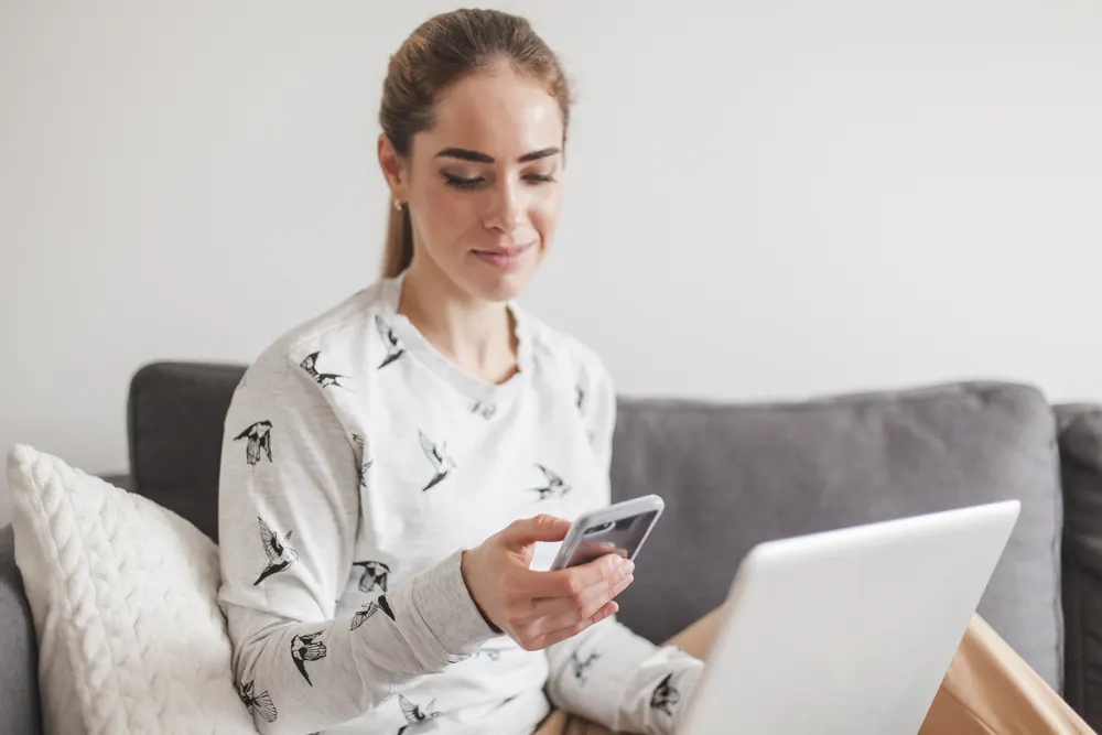 Frau sitzt mit Smartphone und Laptop auf dem Sofa